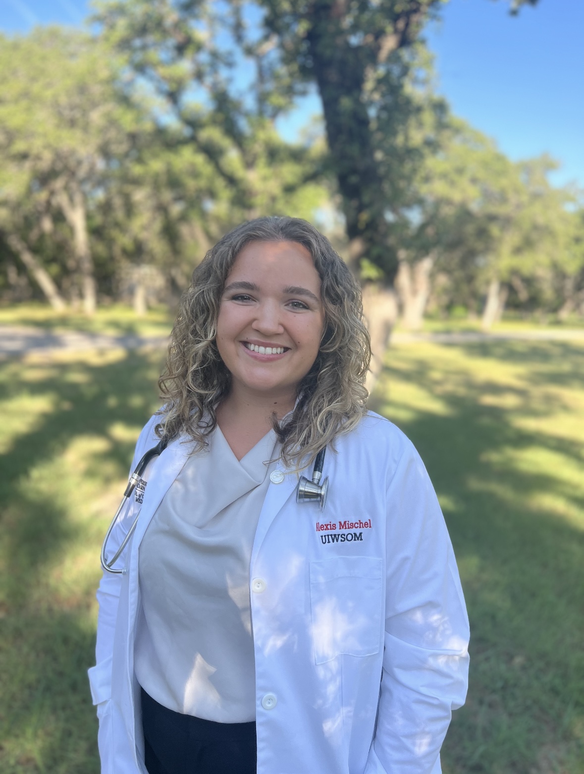 Alexis Mischel is standing under the shade of tall trees on a sunny day in her white coat, smiling at the camera
