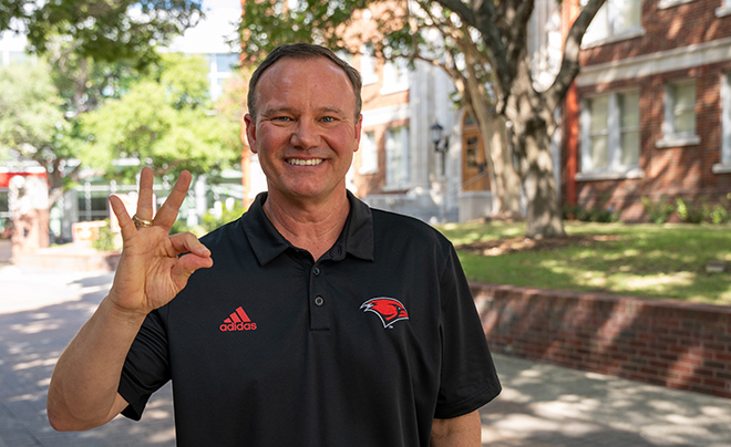 UIW President Thomas Evans standing in corridor of Broadway campus with fingers in Cardinal sign